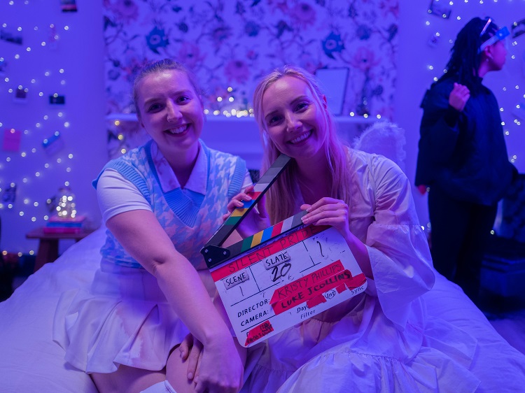 two people sitting on a bed on a film set holding a clapperboard. A member of crew is standing in the background