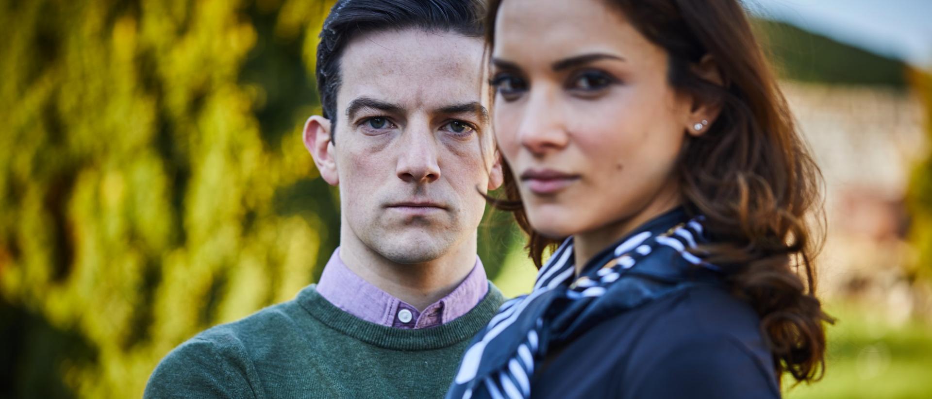 a man and woman stand in a garden looking serious