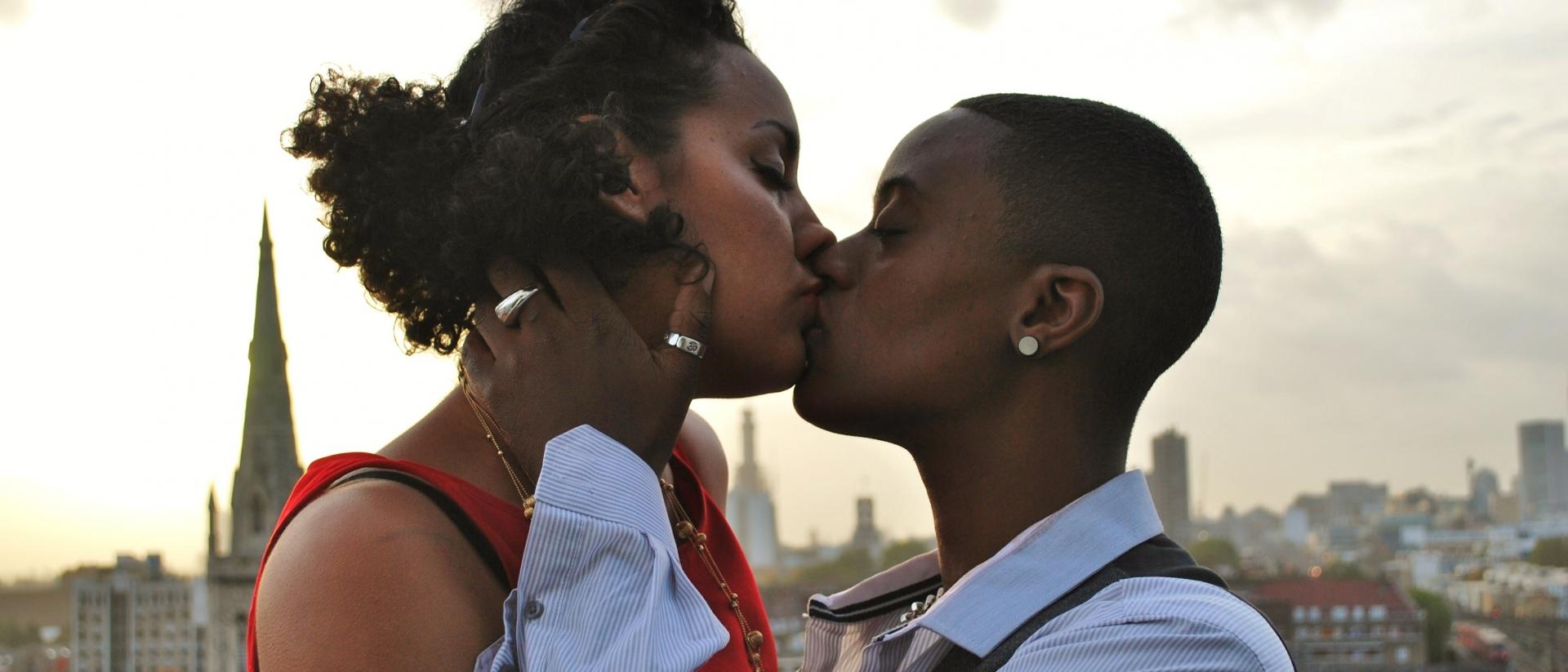 a still from queerama featuring two people kissing on a roof overlooking a city