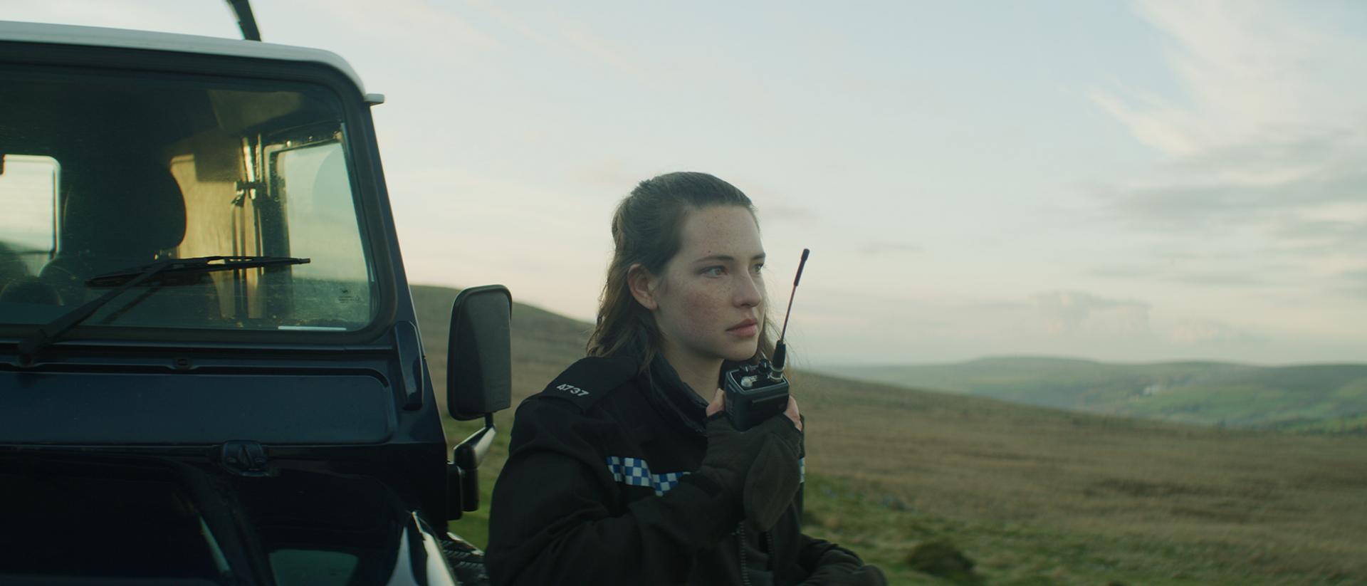 a still from the toll featuring annes elwy as a police officer standing outside next to a jeep. She is talking into a radio.