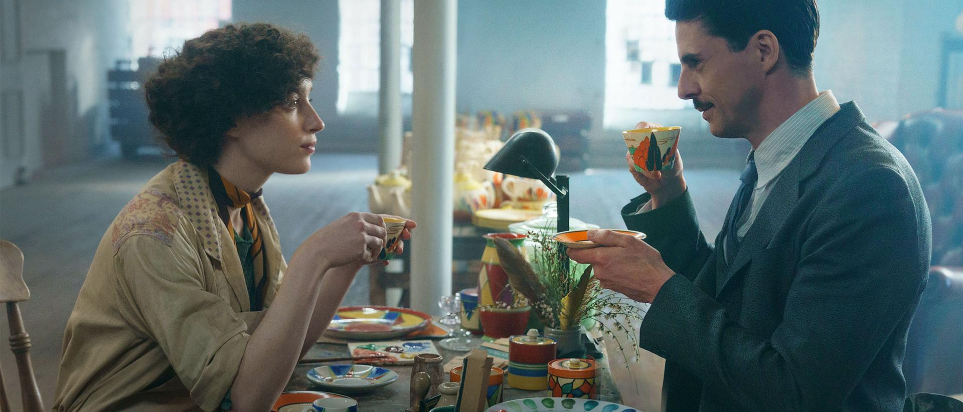 a still from The Colour Room featuring Phoebe Dynevor and Matthew Goode sitting at a table full of ceramic plates, teacups and saucers. They are both holding teacups up to drink from.