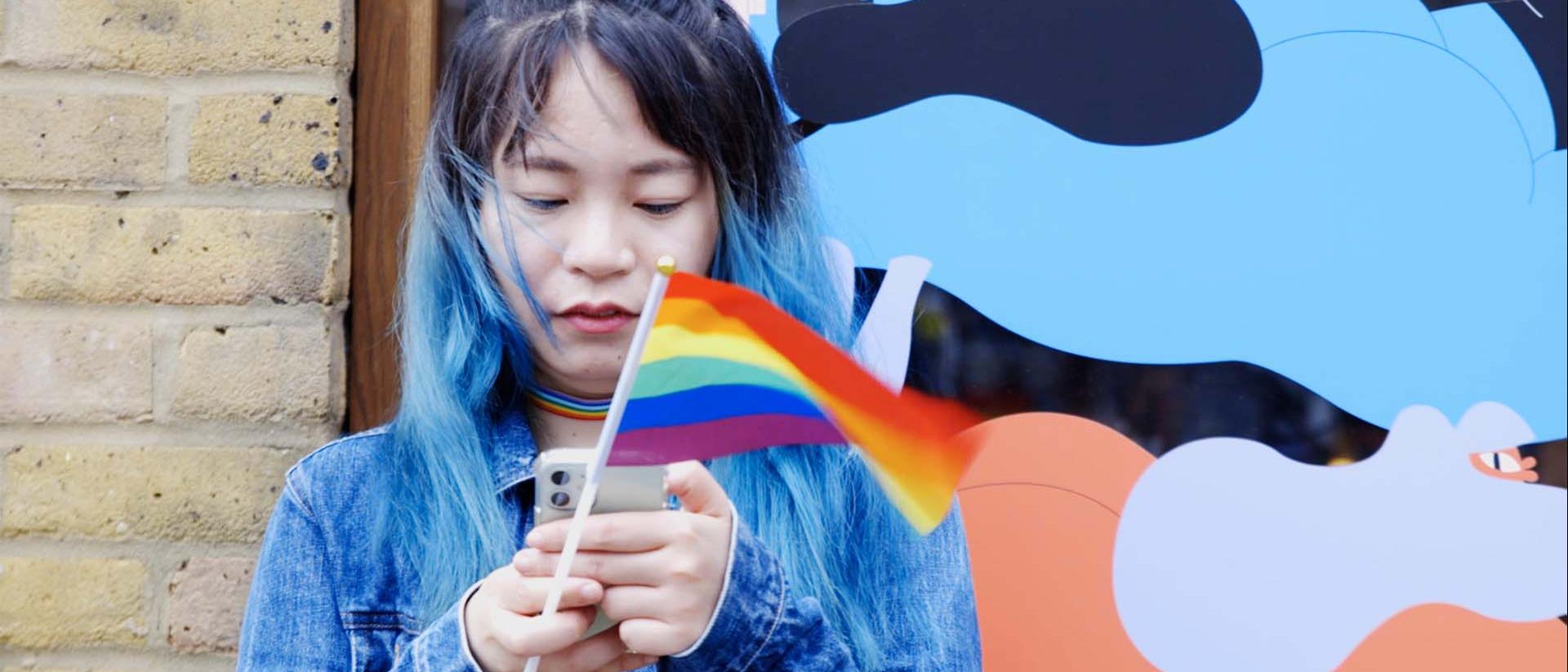 a photo of a person standing outside looking at their phone wile holding a small rainbow flag