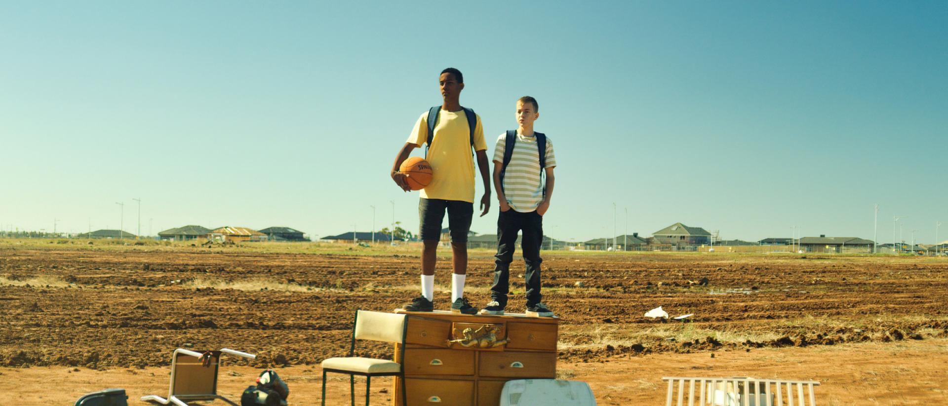 two teenagers stand on an old chest of drawers in an empty field.