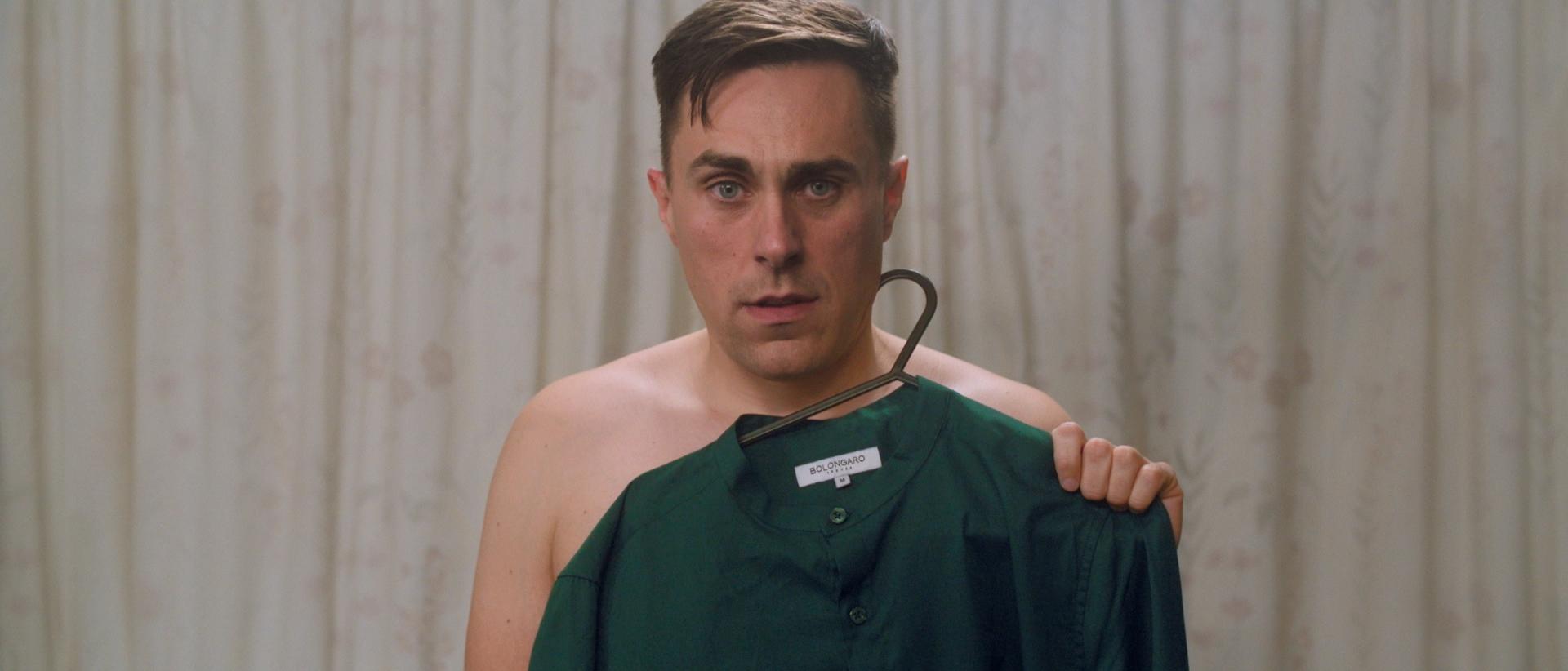 a man stands in a changing room holding a dark green shirt over his torso and staring into camera