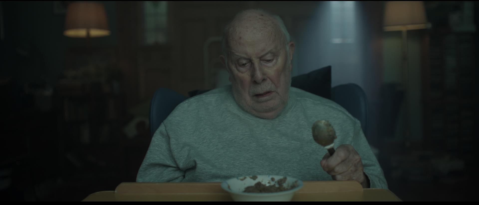 an elderly man sitting in a hospital chair looking down at a bowl of porridge