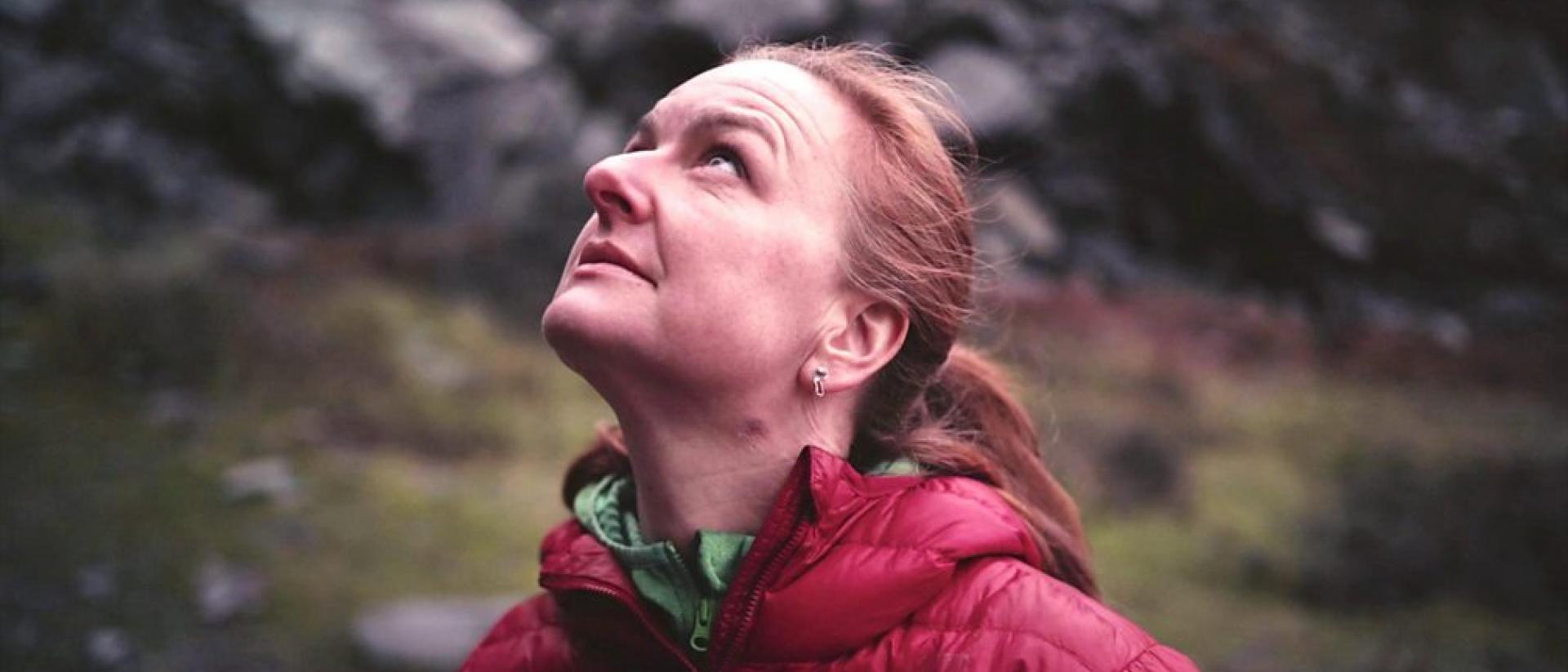 Still from Echdoe featuring a person wearing a red coat looking up. There is a rocky mountainside behind them.