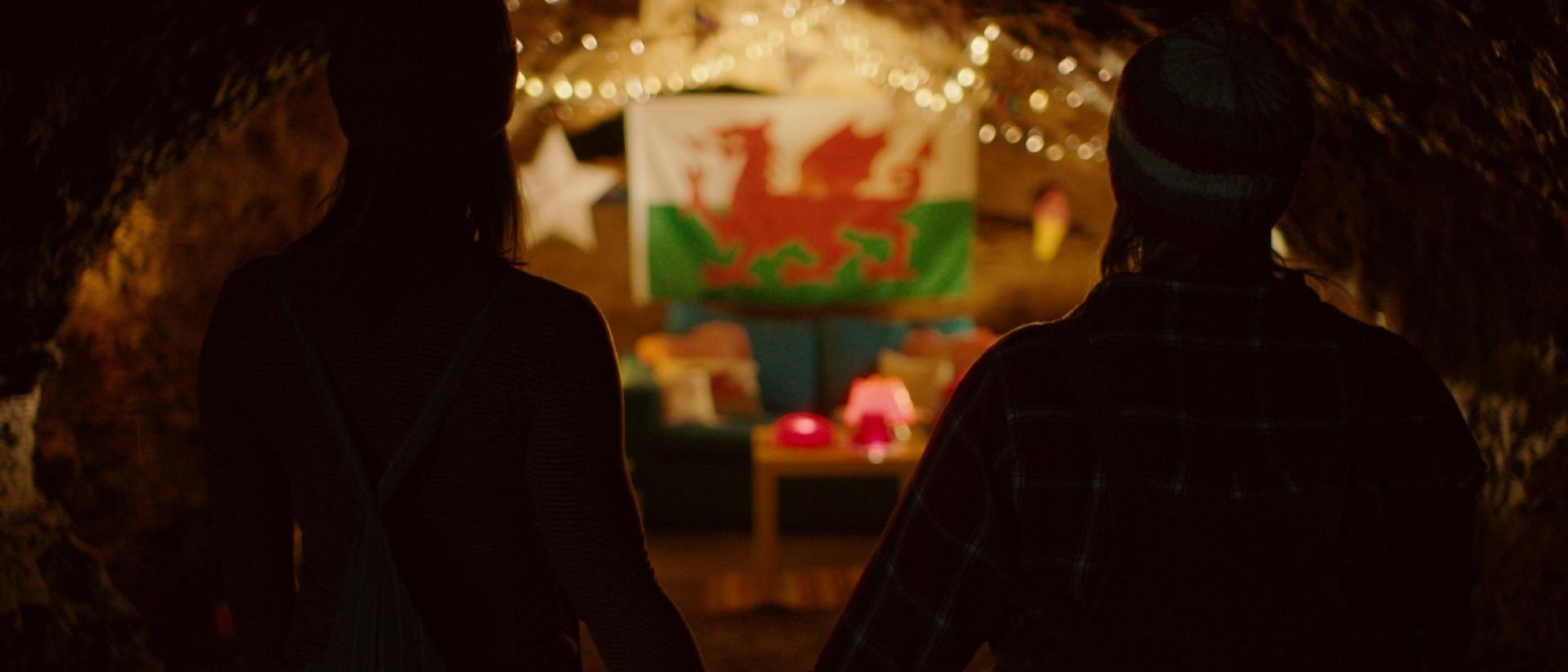 still from jelly featuring two people standing in a cave and holding hands while they look at a welsh flag on the wall, decorated with fairy lights