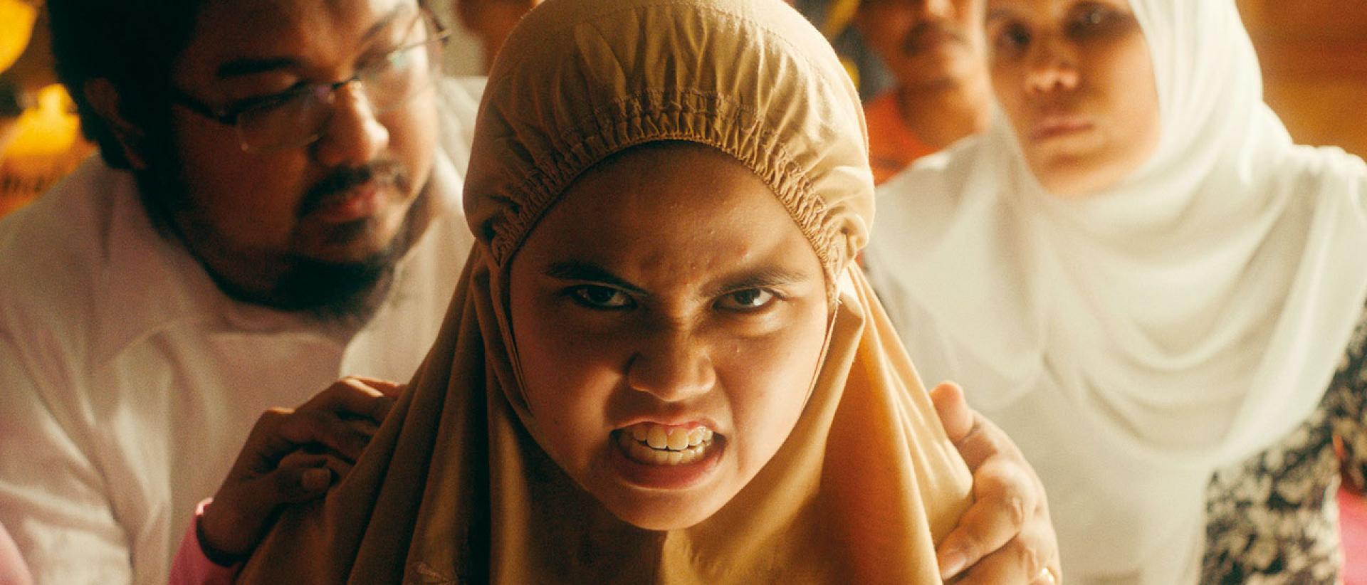 A close-up of a young girl wearing a beige, tudung-style head scarf, an intense snarl on her face. Behind her a man is holding her shoulders and beyond him other adults look on.