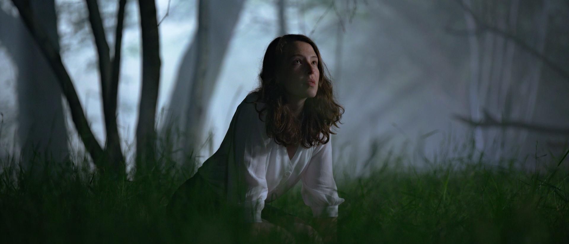 still from gwledd featuring annes elwy wearing a white shirt kneeling in a dark forest