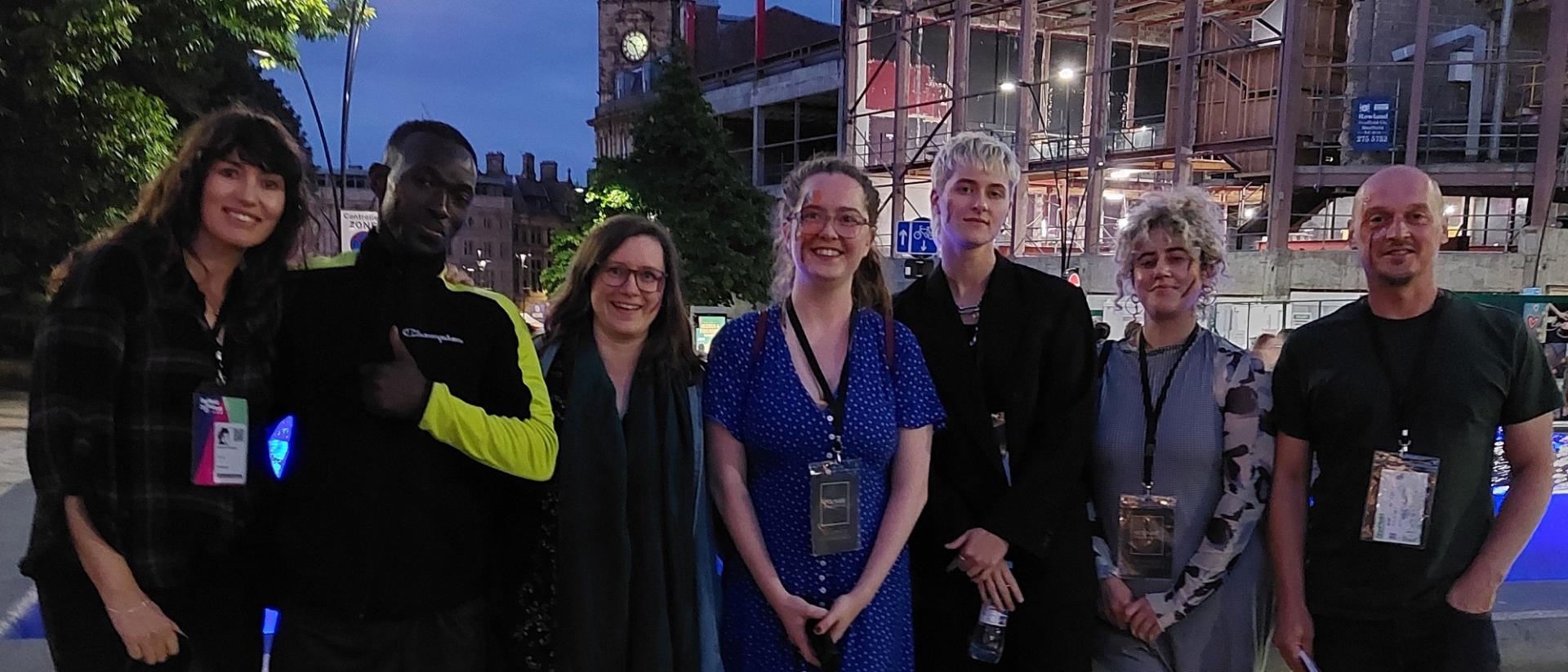seven people standing in a line posing for a photo outside in the evening. there are buildings, including a clock tower, and trees behind them