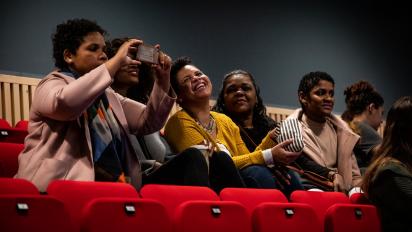 a group of people sitting in a row of cinema seats. They are smiling and laughing. One takes a photo of the cinema screen with her phone.