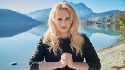 a portrait of rebel wilson standing outside in front of a lake with mountains in the background