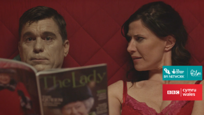 a still from short film stuffed featuring a woman sitting up in bed next to her stuffed dead husband, who is holding a magazine