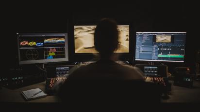 photo of a person sitting in a dark room in front of three computer monitors editing footage.