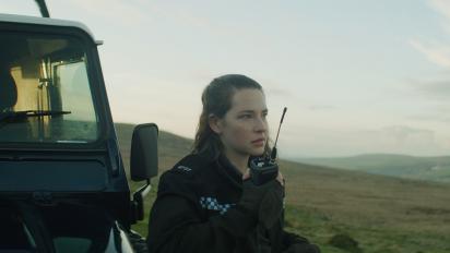 a still from the toll featuring annes elwy as a police officer standing outside next to a jeep. She is talking into a radio.