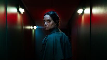 a still from censor featuring Niamh Algar as Enid, standing in a darkened hallway with red walls and looking back towards the camera.