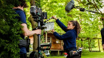 A photo of a film shoot outside a wood cabin in a forest. One person holds a clapperboard up in front of another person with a camera