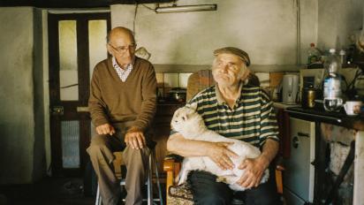 a photo of two elderly men sitting in a farmhouse kitchen. One has a small sheep on his lap.