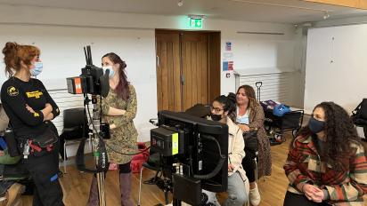 five people behind the scenes of an indoor television set with filming equipment