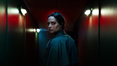 still from censor featuring niamh algar wearing a dark teal coat standing in a dark hallway with red walls and looking back over her shoulder towards camera