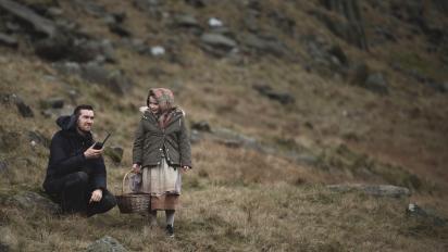 a photo of a child and adult on a film set on a grassy hill. The child is wearing a period costume with a winter coat.