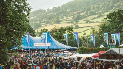 photo from green man festival with lots of people standing in front of a large blue tent. There are trees on a hill in the background.