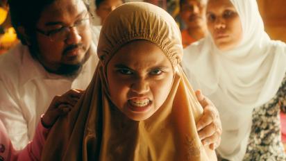 A close-up of a young girl wearing a beige, tudung-style head scarf, an intense snarl on her face. Behind her a man is holding her shoulders and beyond him other adults look on.