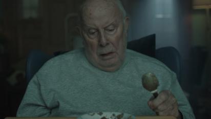 still from g flat featuring an elderly man sitting in a hospital chair, holding a spoon and looking down at a bowl of porridge.