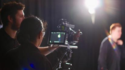 two people using a camera to film another person standing in front of a black curtain