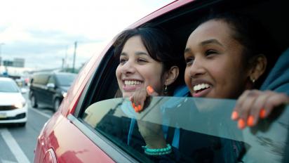 still from brides featuring two teenage girls in the backseat of a car peering out of the half-open window and smiling