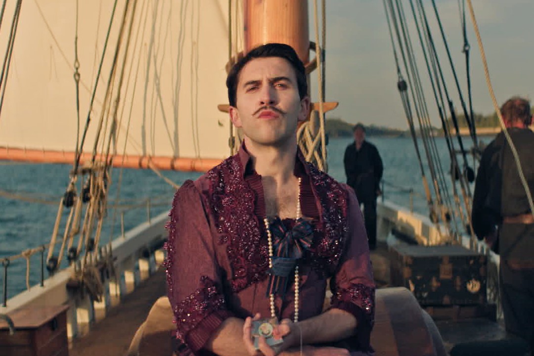 Still from Madfabulous featuring Callum Scott Howells wearing a burgundy 19th century gown and a pearl necklace. They are standing on a sailboat on a body of water with two crew members in the background.