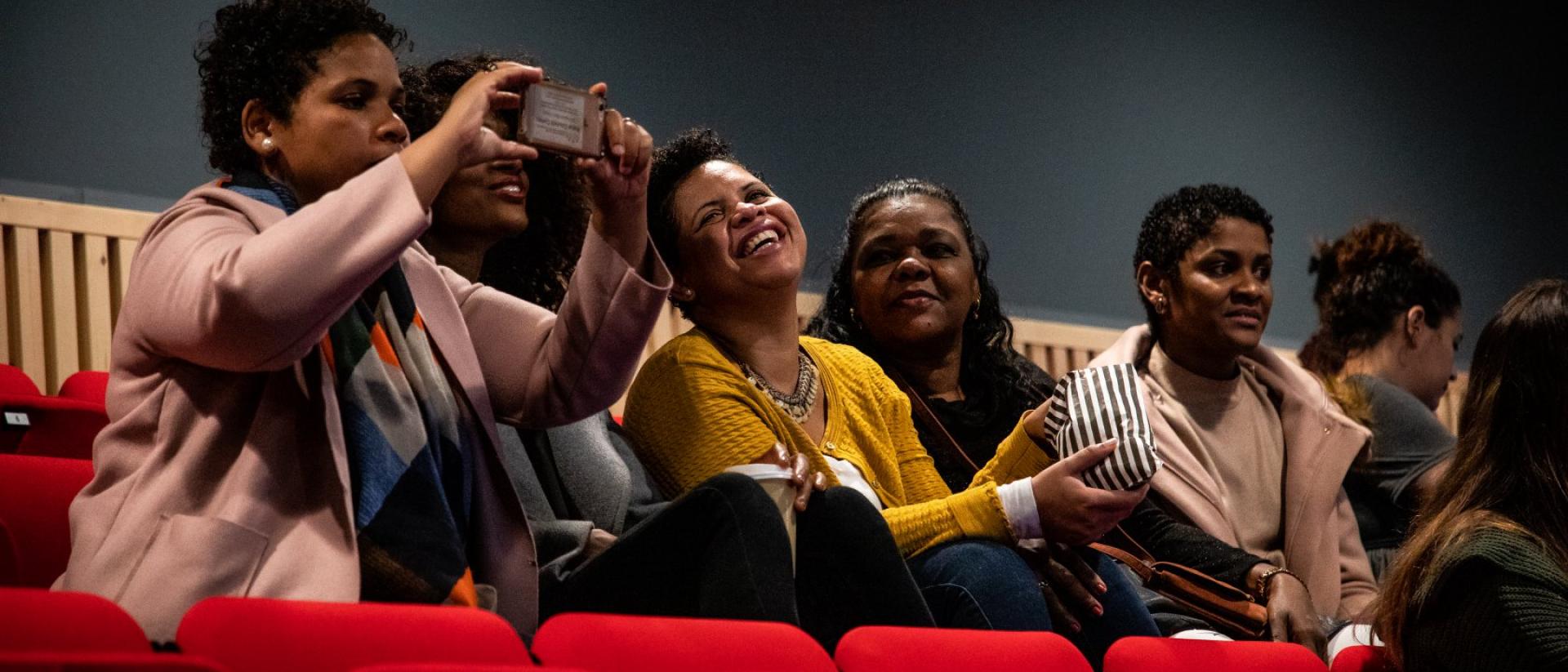 a group of people sitting in a row of cinema seats. They are smiling and laughing. One takes a photo of the cinema screen with her phone.