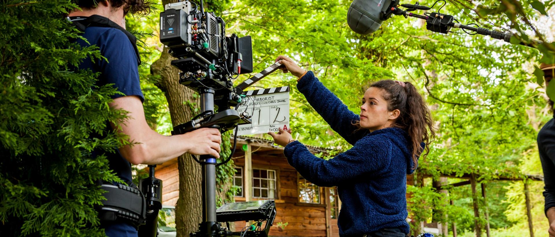 a camera crew shooting in a green forest in front of a wood cabin
