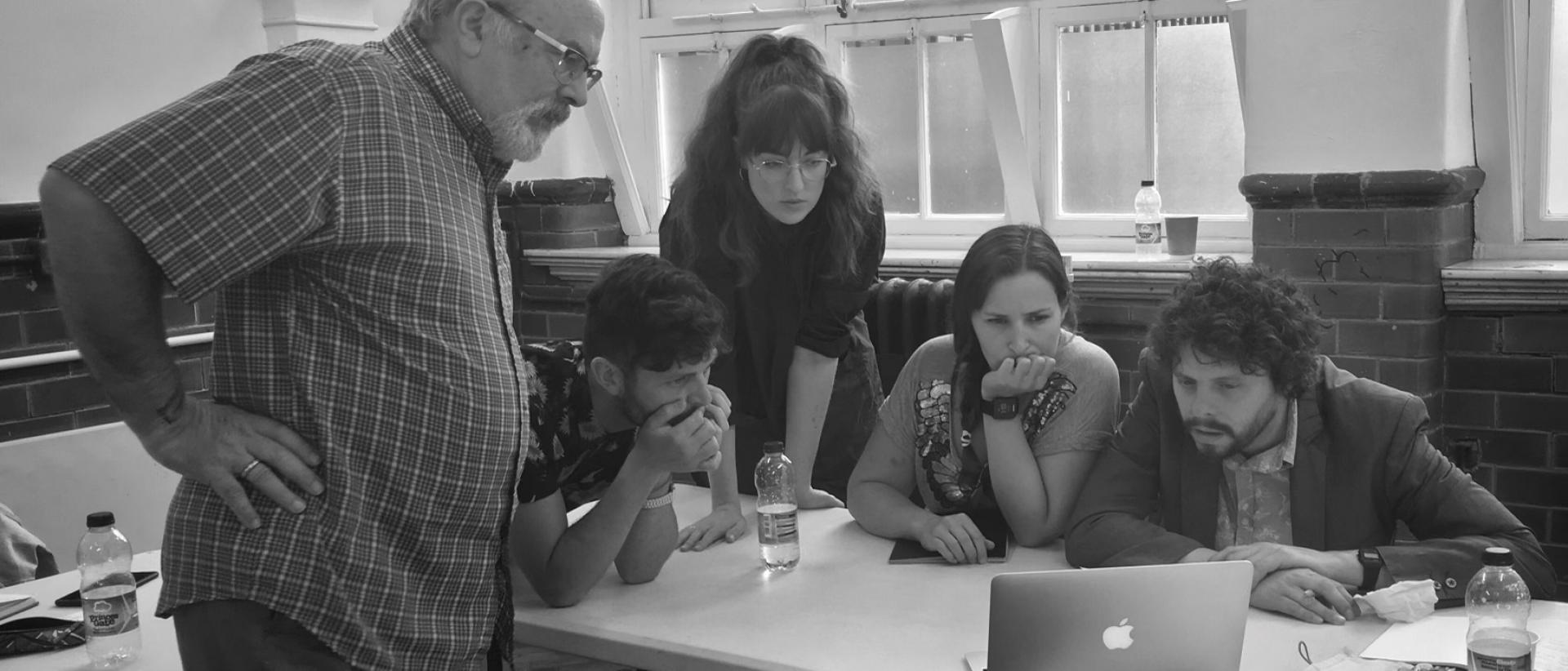 a black and white photo of five people standing and sitting at a desk watching something on a laptop