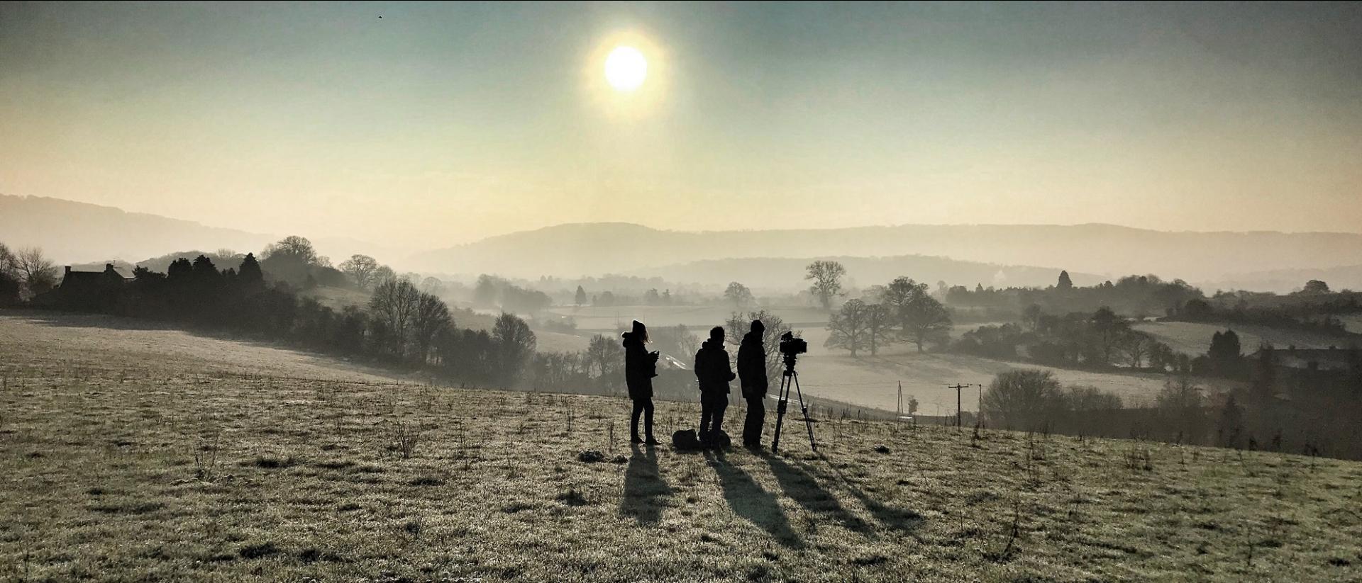 a small film crew standing in a field overlooking a misty valley