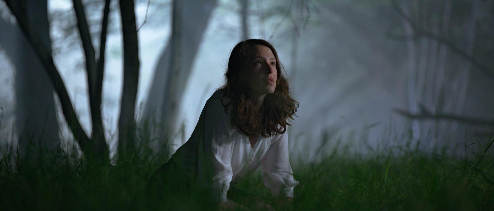 still from glwedd featuring annes elwy wearing a white shirt kneeling down in a dark forest