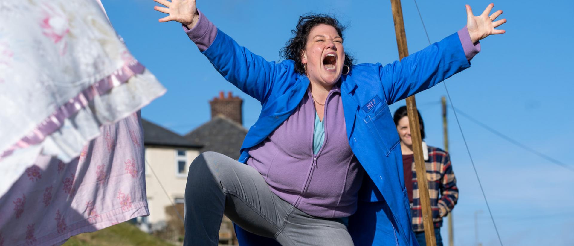 still from chuck chuck baby featuring a woman wearing a blue factory coat standing in front of a washing line and singing with her arms outstretched 