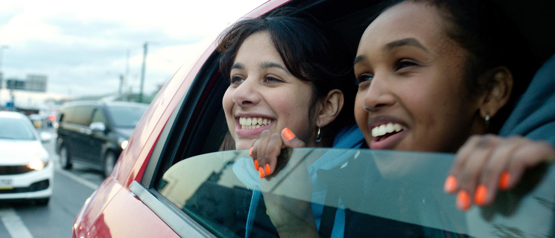 still from brides featuring two teenage girls in the backseat of a car peering out of the half-open window and smiling