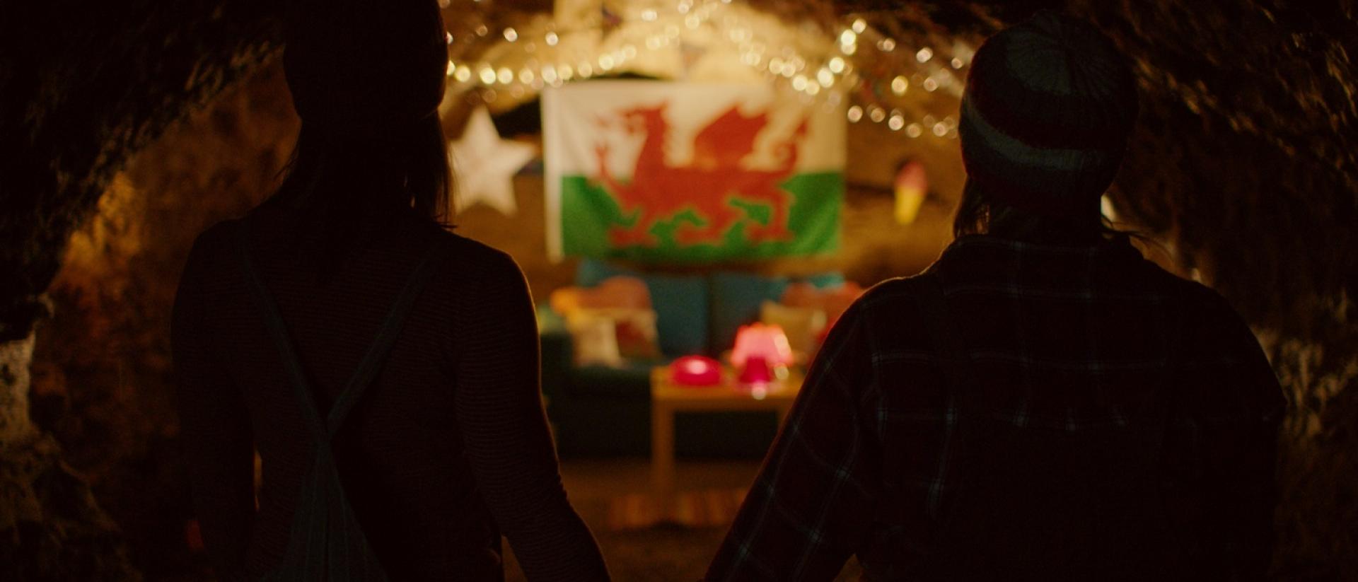 still from jelly featuring two people standing in a cave and holding hands while they look at a welsh flag decorated with fairy lights