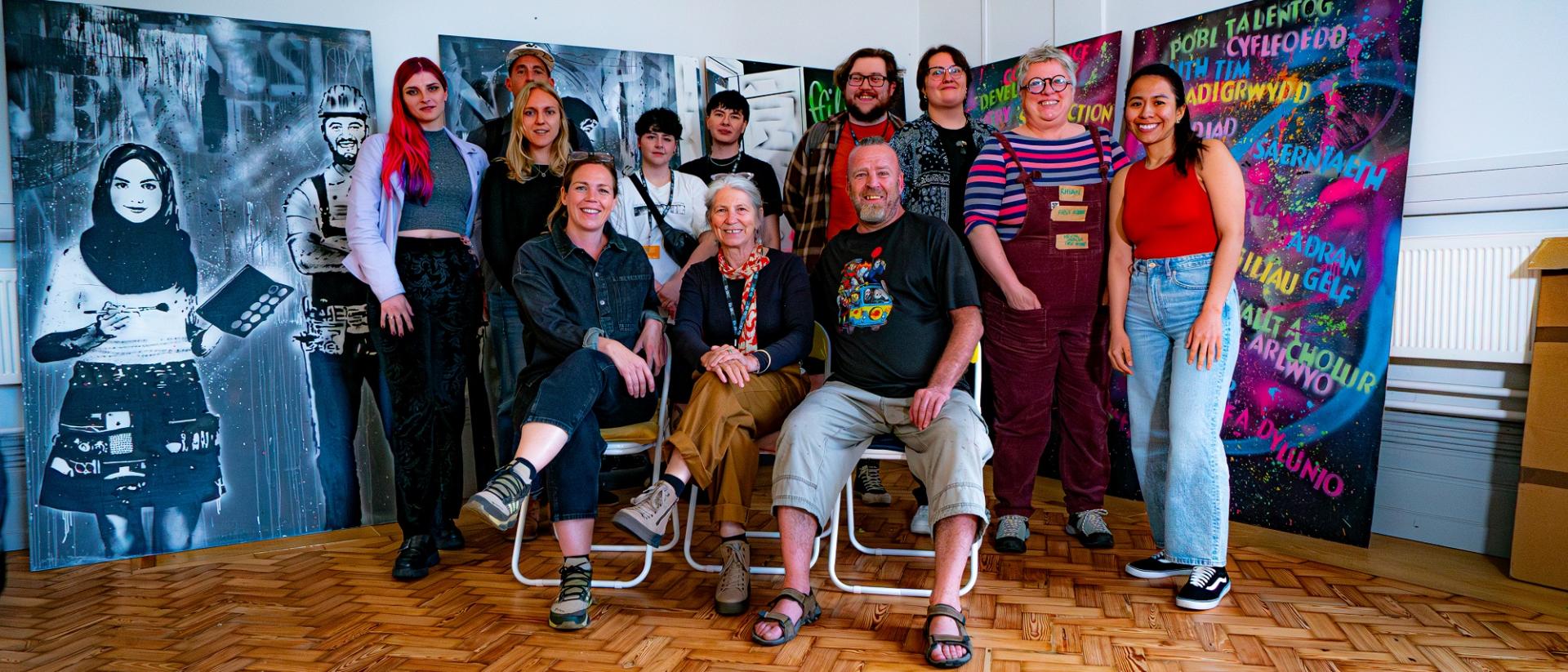 12 people posing for a photo in front of a mural. three are sitting in chairs at the front