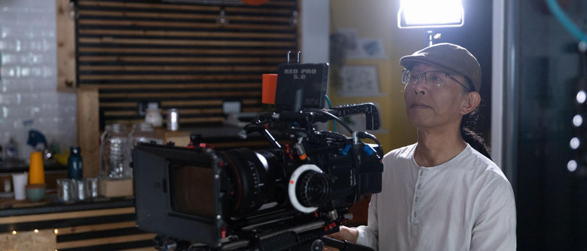 Photo of Keefa Chan standing in a community arts space, operating a camera a on a tripod with a light behind him. He is wearing a white top, a brown flat cap and glasses.