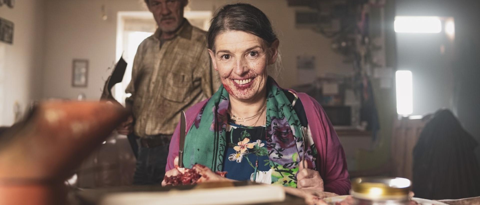 a woman sitting at a farmhouse kitchen table grinning creepily at the camera. A man stands behind her,