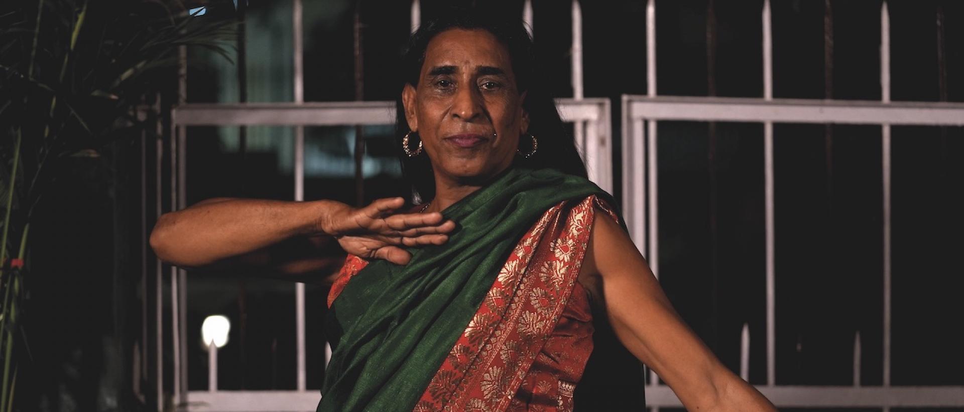 Still from India's First Best Trans Model Agency featuring a person wearing a green and red sari and posing for a photo in front of some metal gates.