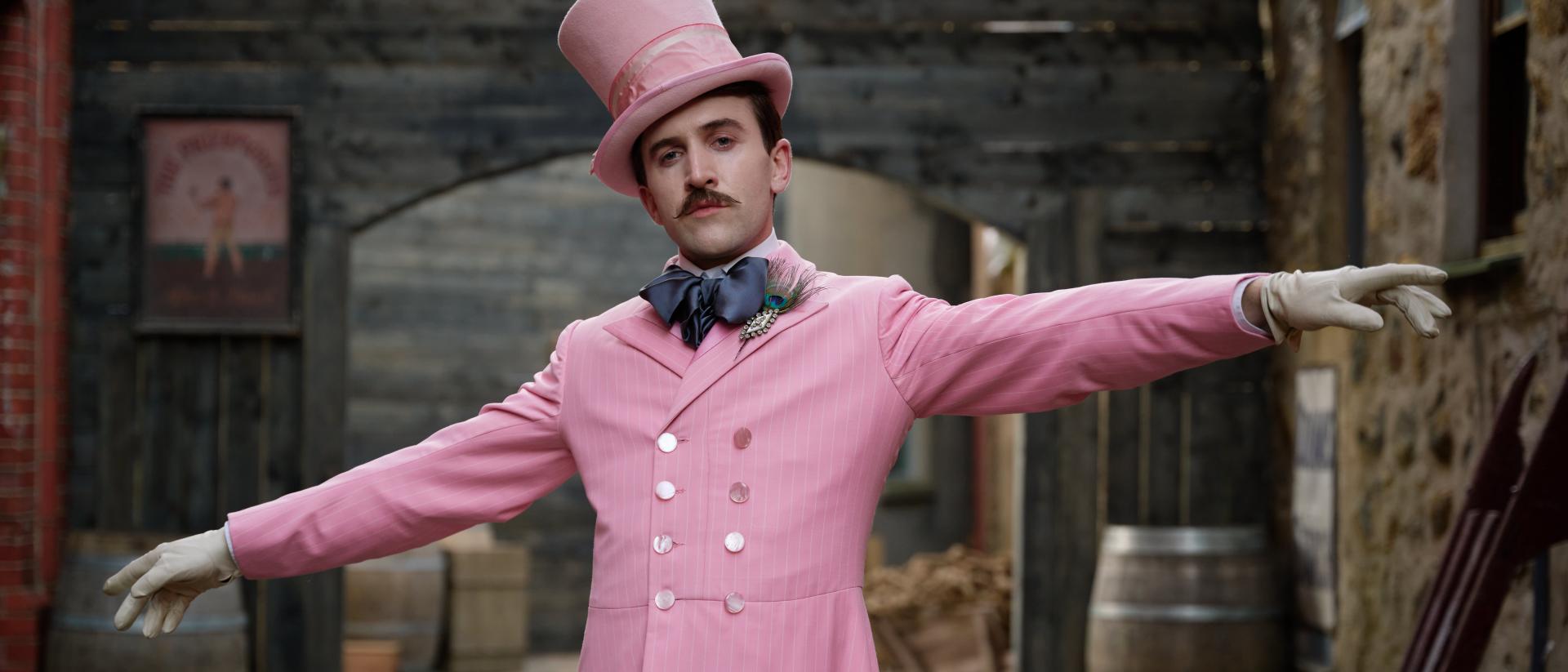 Still from Madfabulous featuring Callum Scott Howells wearing a pink suit and top hat and standing in a 19th century street. There are wooden barrels in the background.