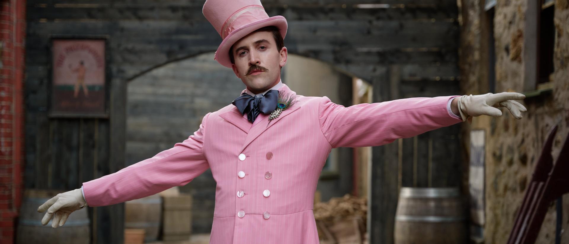 Still from Madfabulous featuring Callum Scott Howells wearing a pink suit and top hat and standing in a 19th century street. There are wooden barrels in the background.