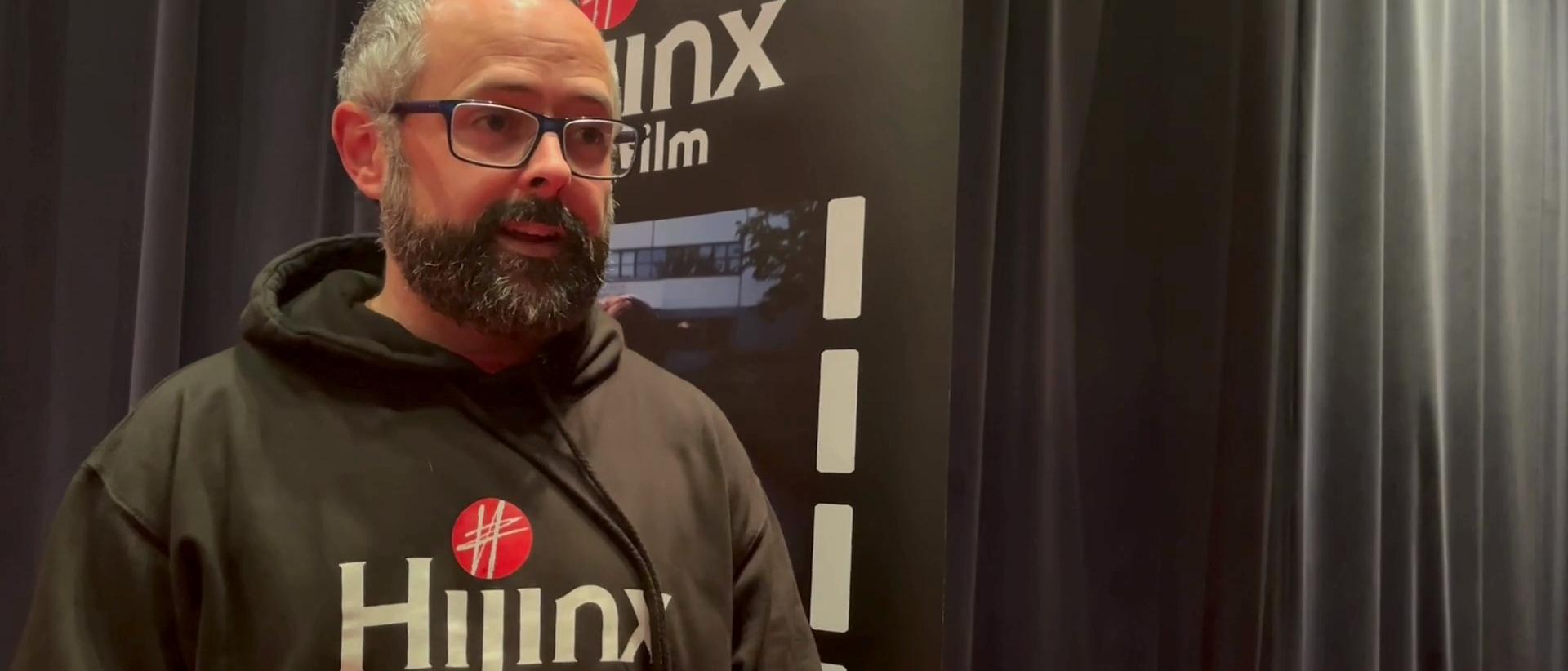 dan mcgowan wearing a black hoodie with Hijinx printed on it, standing in a small cinema screen in front of a Hijinx banner