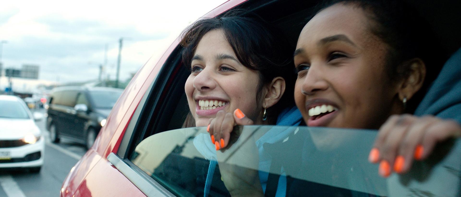Still from Brides featuring two young women looking out of a half-open car window and smiling.