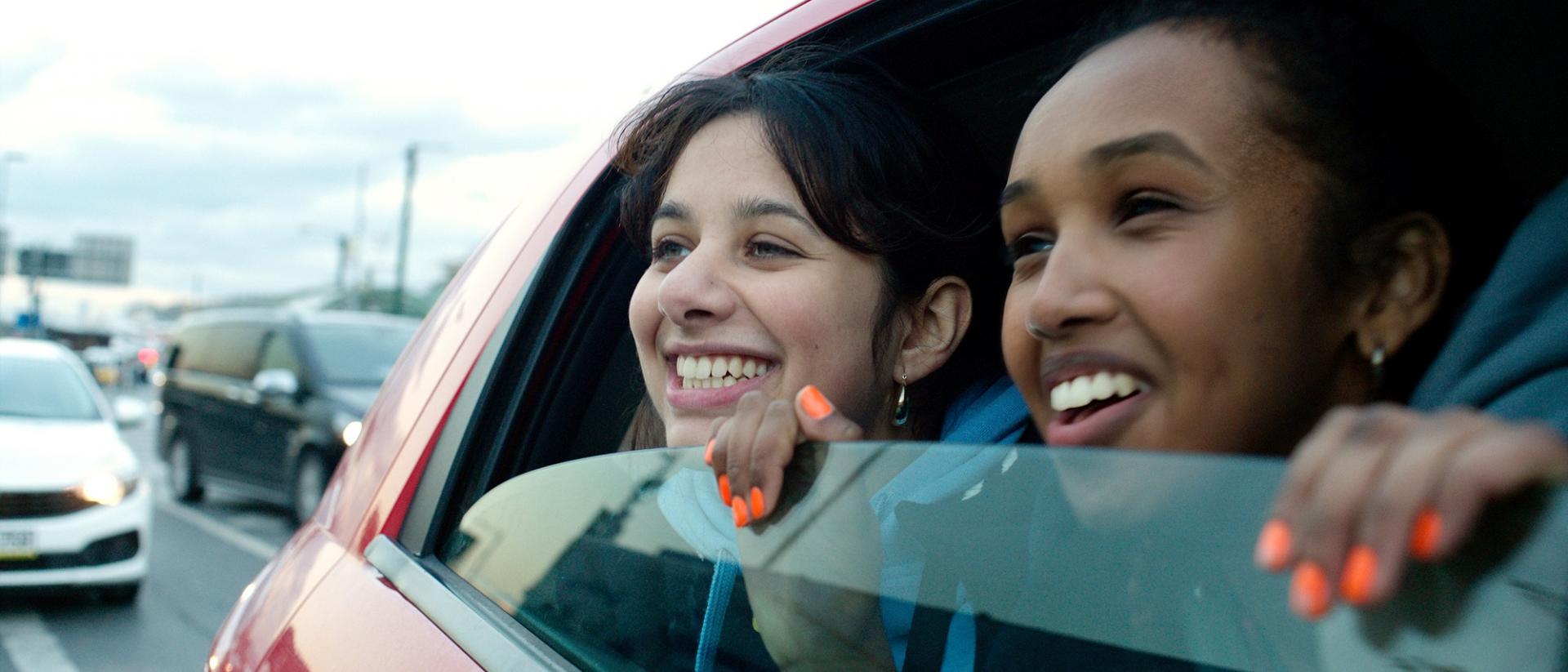 still from brides featuring two teenage girls in the backseat of a car peering out of the half-open window and smiling