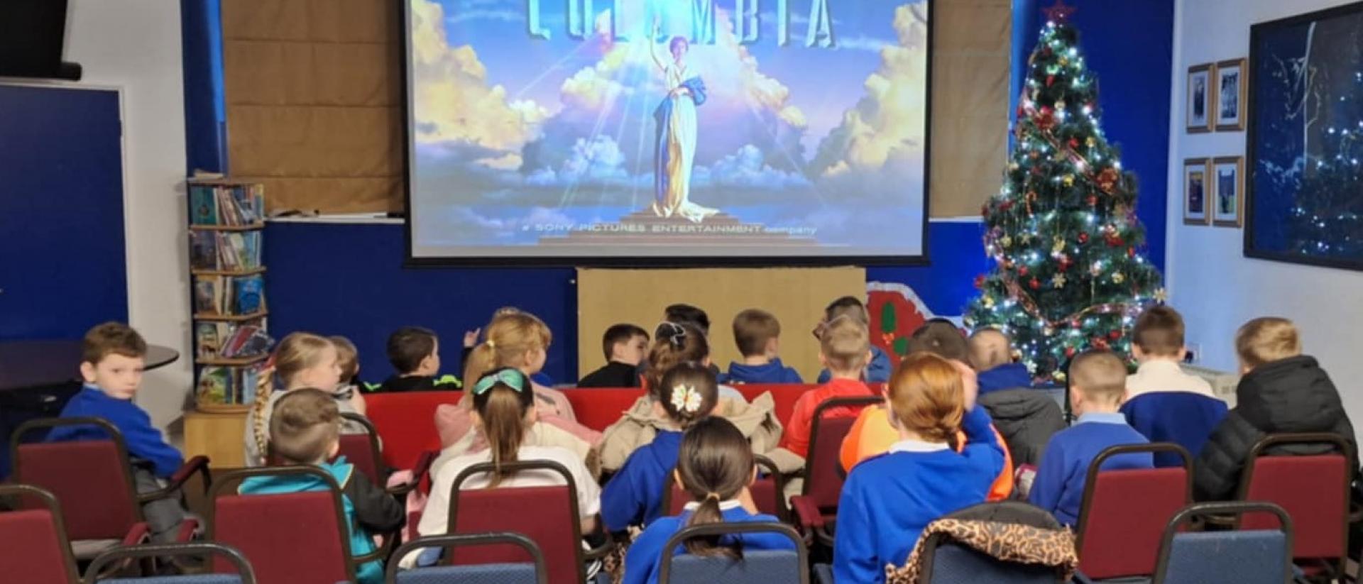 photo of children in blue school uniforms watching a film at a pop-up community cinema. The Columbia logo is on the projector screen and there is a Christmas tree to its right.
