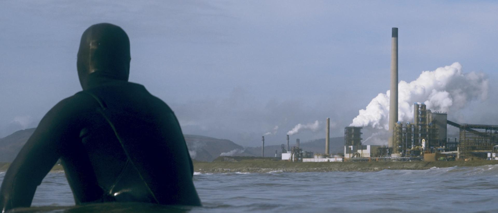 still from documentary smoking shores featuring a surfer in a wetsuit floating in the sea and looking at a steel factory in the background.
